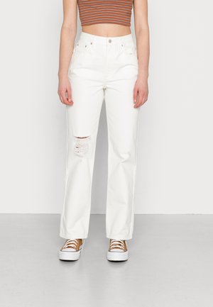 Jeans brancos de perna reta com detalhes desgastados no joelho, combinados com ténis castanhos e brancos. A parte de cima é uma t-shirt cropped de riscas multicoloridas.