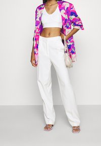 Mujer con top corto blanco, pantalones blancos de cintura alta, blazer floral en tonos rosa y púrpura, sandalias lavanda y un pequeño bolso beige de hombro.