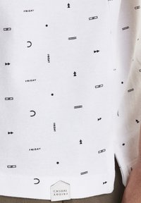 Bílá bavlněná košile s geometrickým vzorem v černé barvě, do kterého je zakomponováno slovo "PÁTEK". Štítek viditelný na lemu uvádí "CASUAL FRIDAY."