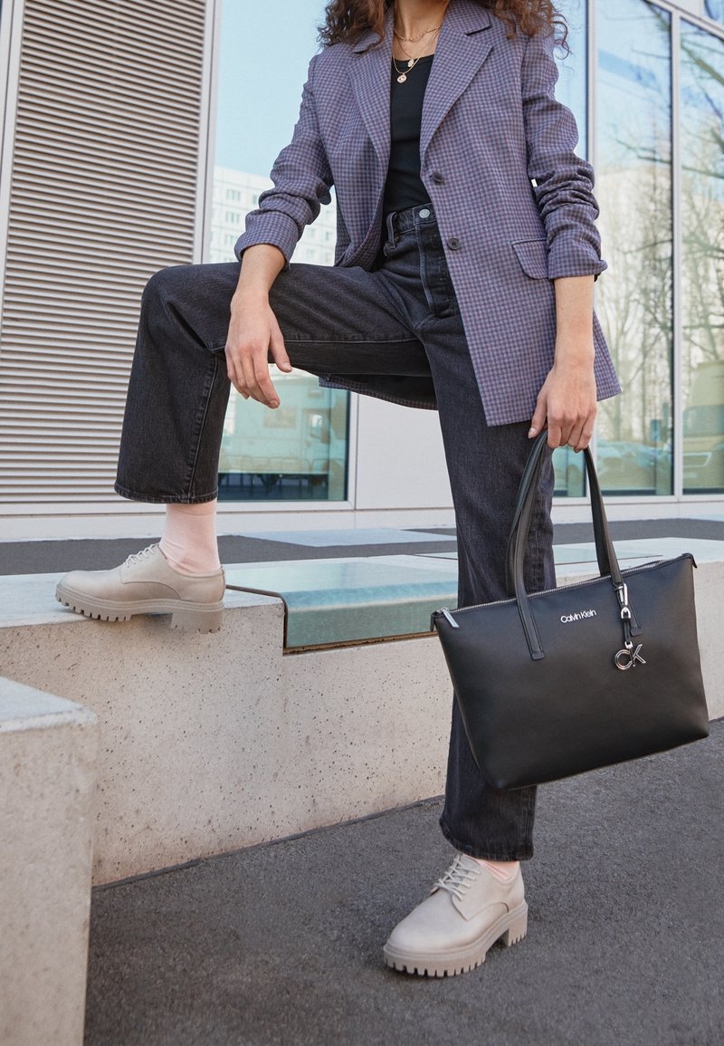 Sac fourre-tout en cuir noir avec matériel en argent, blazer à carreaux, jean noir, et chaussures à plateforme gris clair avec semelle épaisse et chaussettes roses.