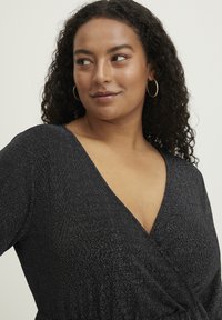 Schwarzes Kleid mit tiefem V-Ausschnitt, gefertig aus strukturiertem Stoff mit silbernen Fadenmustern, sanfter Drapierung und langen Ärmeln.