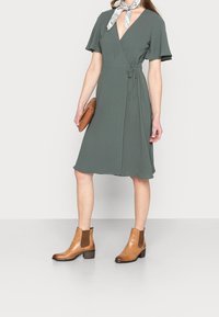 Vihreä kietaisumekko, jossa on liehuvat hihat, vyötärölle solmittava yksityiskohta ja V-pääntie. Yhdistetty ruskeisiin nilkkabootseihin, joissa on elastiset sivupaneelit.