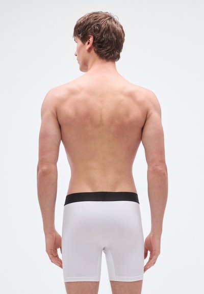 Boxer de algodón blanco con una cintura elástica negra. El modelo está de pie de espaldas, mostrando el ajuste y el diseño de los shorts.