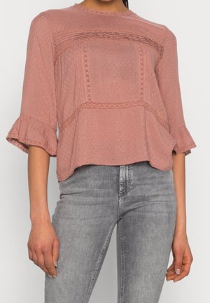 Femme portant un chemisier texturé rose poudré avec des détails en dentelle et volants, associé à un jean gris clair délavé.