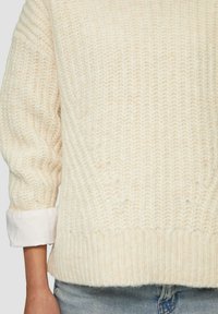 Crèmekleurige gebreide trui met een gestructureerd patroon en een geribbelde zoom. Heeft zichtbare naden en opgerolde mouwen over een licht shirt.