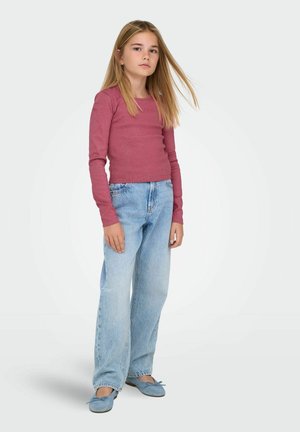 Jeune fille aux cheveux blonds portant un haut rose à manches longues, un jean bleu clair ample et des ballerines bleues sur un fond uni.