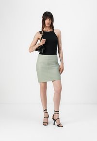 Svart, ermeløs topp kombinert med et tettsittende lys grønt skinnskjørt. Modellen har på seg svarte klaffehæler og holder en liten svart håndveske.