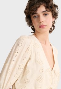 Cremfarbene Bluse mit V-Ausschnitt, puffartigen Ärmeln, floralem Ausschnitt-Detail und Schleife vorne, aus leichtem strukturiertem Stoff gefertigt.