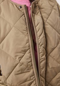 Veste matelassée beige avec boutons-pression et fermeture éclair, portée ouverte sur un pull rose, laissant apparaître une partie de longs cheveux foncés.
