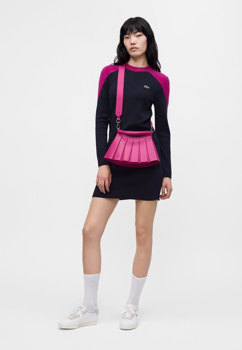 Femme portant une robe bleu marine avec des épaules roses, des chaussettes blanches, des baskets blanches et un sac à bandoulière plissé rose vif, debout devant un fond uni.