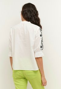 Femme aux longs cheveux bouclés portant un chemisier blanc avec une broderie florale noire sur les manches et un pantalon vert citron, vue de dos.