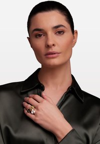 Anello floreale con più colori, caratterizzato da petali bianchi, gialli e marroni, montato su una fascia dorata. Il modello indossa una camicia di satin scuro.