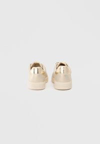 Et par beige sneakers med metallisk gullhæl, sett fra baksiden mot en enkel hvit bakgrunn.