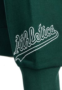 Tessuto a maglia verde con la parola "Athletics" ricamata in bianco e un dettaglio di sottolineatura curvata vicino a un polsino a coste.