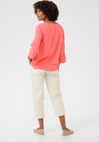 Blusa corallo con maniche a tre quarti, presenta un collo tondo e una piega sul retro; abbinata a pantaloni cropped beige chiaro e slip-on beige.