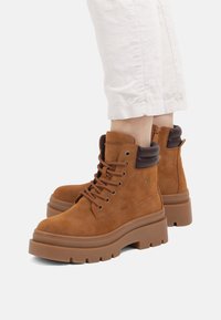 Botas de tobillo de ante marrón con suela gruesa, diseño de cordones y acentos en marrón oscuro contrastante en el cuello. Presentan un detalle de logo.