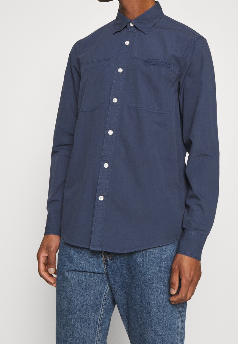 Homme portant une chemise bleu marine boutonnée avec des poches sur la poitrine à l’avant et un jean bleu, debout devant un fond clair uni.