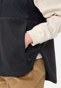 Schwarze Fleece-Weste mit glattem oberen Abschnitt, Seitentaschen-Details und einem kontrastierenden hellen Ärmel darunter.