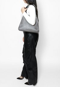 Borsa a mano grigia intrecciata con un design testurizzato e manici neri, tenuta da una persona che indossa un top bianco e pantaloni neri.