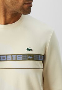Mann som har på seg en beige sweatshirt med grønn og blå horisontale striper, med en liten grønn krokodille-logo på brystet.