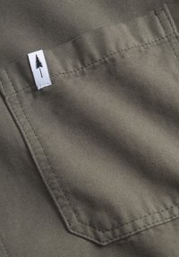 Tela verde con una superficie texturizada, que presenta un bolsillo cosido y una etiqueta blanca con un logotipo de árbol negro.