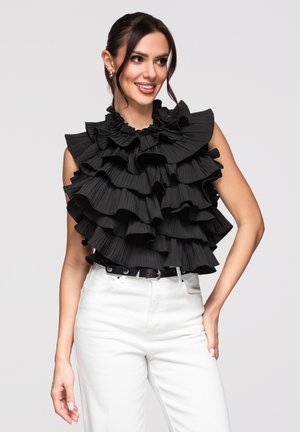 Kobieta nosząca czarny top bez rękawów z wielowarstwowymi falbanami, w połączeniu z białymi spodniami z wysokim stanem i czarnym paskiem, uśmiechająca się.