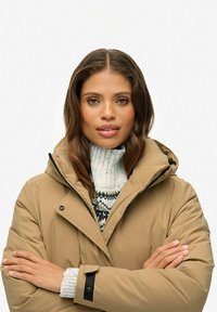 Beige geïsoleerde jas met capuchon, ritssluiting en zwarte accenten, gedragen over een chunky, lichtgekleurde gebreide trui met patronen.