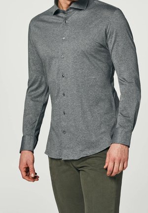 Uomo che indossa una camicia grigia aderente a maniche lunghe con bottoni e pantaloni verde scuro su uno sfondo semplice.