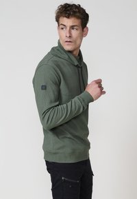 Hombre con un pulóver verde texturizado de alto cuello y pantalones cargo negros, mirando a su izquierda contra un fondo plano.