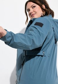 Chaqueta azul impermeable con cierre de cremallera, cinturón ajustable, capucha y tejido texturizado. Incluye un parche negro con el logo en la manga.