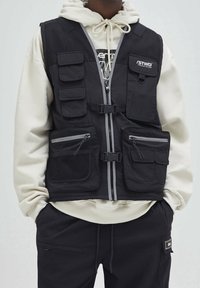 Czarna kamizelka użytkowa z wieloma kieszeniami, panelem z siateczki, zamkiem błyskawicznym z przodu i regulowanymi klamrami. Wykonana z trwałej tkaniny z szarymi akcentami.
