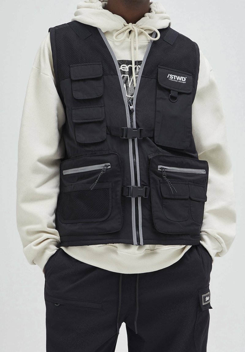 Czarna kamizelka użytkowa z wieloma kieszeniami, panelem z siateczki, zamkiem błyskawicznym z przodu i regulowanymi klamrami. Wykonana z trwałej tkaniny z szarymi akcentami.