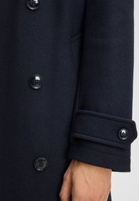 Donker marineblauwe jasje met knopenmanchetten en bijpassende knopen aan de voorkant, waarbij een deel van een hand zichtbaar is.