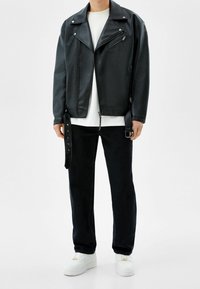 Person som bär en svart läderbikerjacka, vit t-shirt, svarta jeans och vita sneakers stående mot en vit bakgrund.