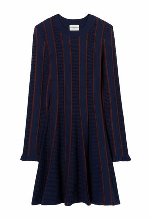 Mujer de pie de frente llevando un vestido azul marino con rayas rojas, zapatos marrones, bolso cruzado y sosteniendo una chaqueta marrón en una mano.