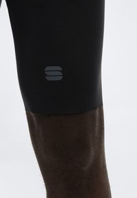 Κοντινό πλάνο ενός σκουρόχρωμου ποδιού που φοράει μαύρα κοντά κολάν συμπίεσης με ένα μικρό αντανακλαστικό λογότυπο κοντά στον μηρό, σε απλό φόντο.