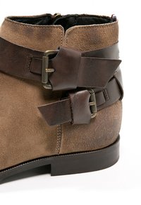 Stivale alla caviglia in camoscio marrone con cinturini in pelle e fibbie in ottone. Finitura testurizzata e punta arrotondata su una suola nera solida.