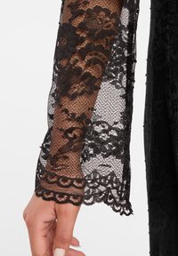 Zwarte kanten mouw met bloemenpatroon en golvende afwerking. Doorzichtige textuur over een effen zwarte kledingstuk. Beschikt over ingewikkelde kantdetails.