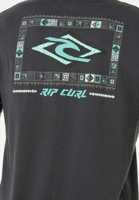 Zwarte katoenen T-shirt met een turquoise golflogo binnen een frame met patronen en geometrische accenten. Tekst leest "RIP CURL."