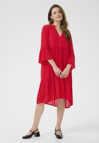 Robe rouge fluide avec un décolleté en V et des manches évasées. Présente des couches à volants et un ourlet légèrement asymétrique. Tissu léger et transparent.