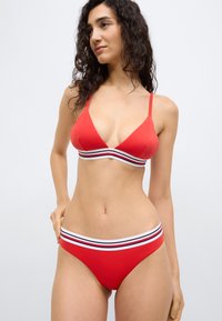 Σετ κόκκινου μπικίνι με τοπ τριγωνικό και σλιπ χαμηλής μέσης. Και τα δύο κομμάτια έχουν ριγέ λευκή και μαύρη ταινία. Λείο, ελαστικό υλικό.