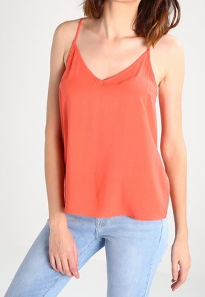 Kobieta ubrana w pomarańczowy top na ramiączkach z dekoltem w serek i jasnoniebieskie dżinsy na tle jednolitego, jasnego tła.