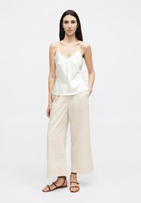 Žena v bílém topu, béžových širokých pruhovaných kalhotách a hnědých sandálech stojí s jednou rukou v kapse.