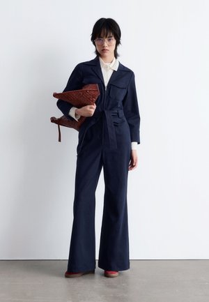 Navy blue jumpsuit with flared legs, two front pockets, and a belted waist. Holding a woven brown bag, paired with red shoes.