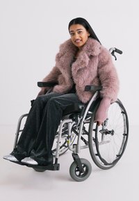 Cappotto in pelliccia sintetica rosa, pantaloni neri glitterati, seduta in una sedia a rotelle con scarpe argentate. Lo sfondo semplice enfatizza la texture e il colore dell'outfit.