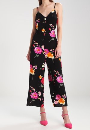 Vrouw draagt een zwarte mouwloze jumpsuit met roze en oranje bloemen en roze hoge hakken, staand tegen een effen achtergrond.