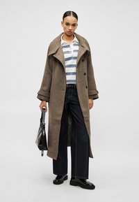 Mulher vestindo um casaco longo castanho sobre uma camisa branca às riscas, jeans escuros de perna larga, sapatos pretos e segurando uma mala preta.