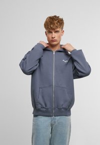 Gray zip-up hoodie with a kangaroo pocket, relaxed fit, and drawstring hood. Features embroidered logo on the chest. Soft fabric texture.