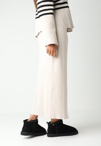 Krémová plisovaná midi sukně v kombinaci s černobílým pruhovaným svetr a černými semišovými kotníkovými botami, zdůrazňující texturované materiály a vrstvený design.