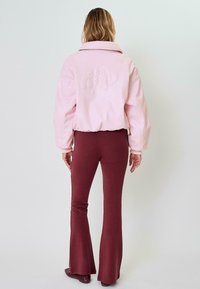 Giacca rosa corta con colletto alto e logo in rilievo sulla schiena, abbinata a pantaloni svasati bordeaux e scarpe coordinate.